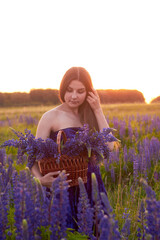 A young girl holds a wicker basket with a large luxurious bouquet of purple lupins, walks across the field at sunset.
