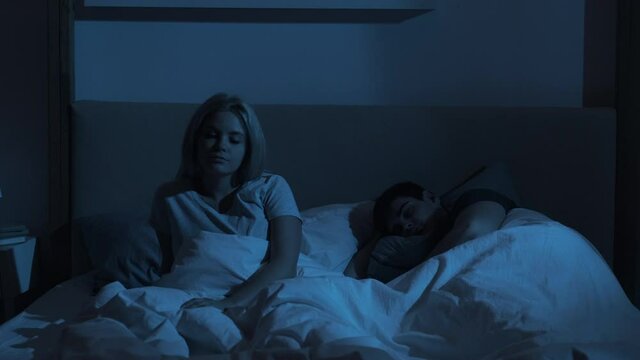 Night insomnia. Sleep disorder. Fatigue anxiety. Exhausted tired bored woman sitting in bed awake with peaceful resting man lying late in dark bedroom with blue light.