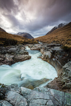 long exposure image of the river etive and waterfalls in glen etive during winter showing flowing water moving over dark rock with scottish mountains in the background