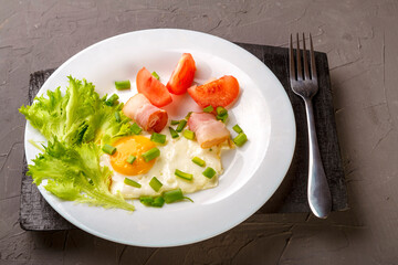 Scrambled eggs with bacon garnished with tomato and green onions on a gray concrete background on a wooden stand next to a fork.