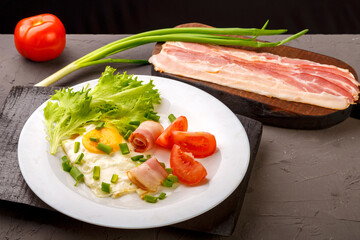 Scrambled eggs with bacon, garnished with tomato and green onion and on a gray concrete background on a wooden stand next to chopped bacon and tomato.