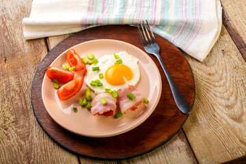 Scrambled eggs with bacon with tomato and green onions and on a wooden table in a plate on a stand with a fork.