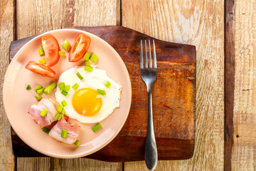 Fried eggs with bacon decorated with tomato and green onion on a wooden board.