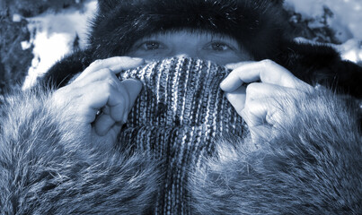 The face of a woman in a winter fur hat and fur coat close-up. Only the smiling, kind eyes are visible, and she covers everything else with a scarf, lifting it up with her hands.
