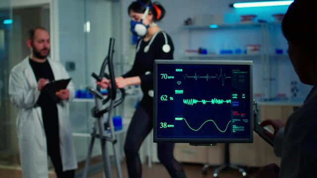 Specialist sport researcher monitoring heart rate on athlete while woman with mask running on cross trainer talking with medical doctor. Physician using tablet controling EKG data in modern laboratory