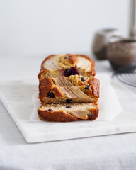 Banana bread, slice of cake with banana and blueberries. Morning breakfast with coffee on light gray linen textile tablecloth on table, Lifestyle still life food. Side view. 