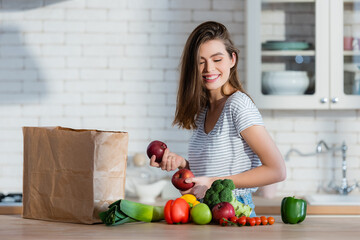 positive woman holding juicy apples near paper bag and vegetables on kitchen table