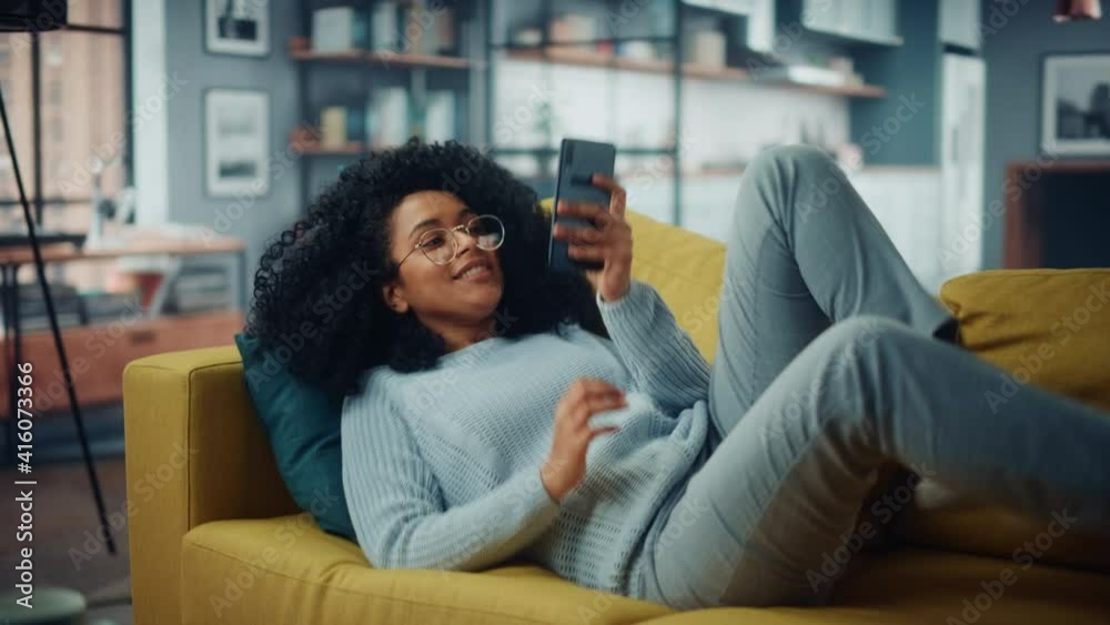 Poster happy young latina with afro hair jumping over on a sofa couch at home while using a smartphone. bea