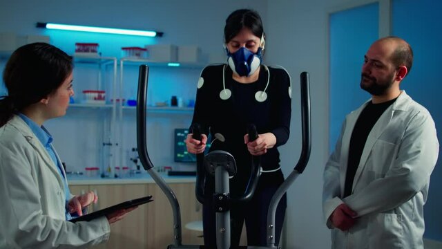 Woman athlete with mask doing fitness exercises in science sport lab with electrodes attached while scientist holding tablet supervising whole process. Physician using notepad controling EKG data - Powered by Adobe