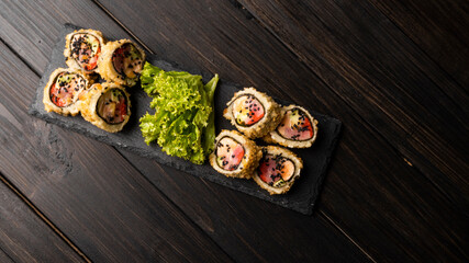 Custom sushi roll in tempura with nori, fresh salmon, tuna, avocado, masago caviar, drizzled with pineapple sauce with salad pouring as decoration on a black plate on a wooden table and background.