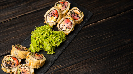 Obraz na płótnie Canvas Custom sushi roll in tempura with nori, fresh salmon, tuna, avocado, masago caviar, drizzled with pineapple sauce with salad pouring as decoration on a black plate on a wooden table and background.