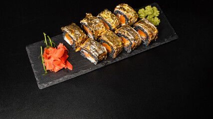 Custom sushi roll with nori, cuttlefish ink, fresh salmon, cream cheese, pepper pumpkin, eel, unagi sauce and edible gold leaf with red ginger, wasabi on a square plate on black table and background.