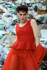 Transgender woman in red clothing, posing, with a landfill garbage background. Lgtb and underground fashion concept.