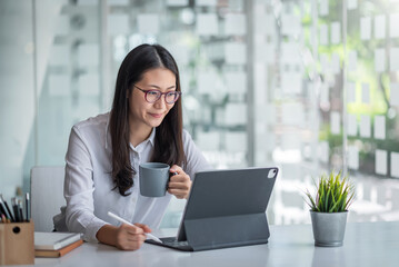 Beautiful smiling Asian woman is happy to work using a tablet and drinking coffee at the office.