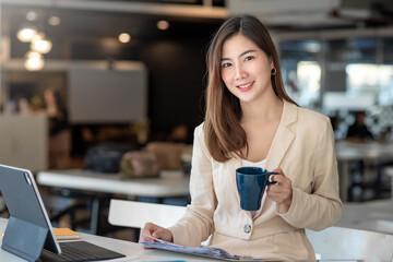Charming Asian woman holding a coffee cup, tablet, document placed on the table at the office. Looking at camera.