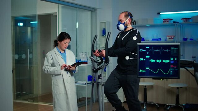 Sportsman with mask running on cross trainer in sports science laboratory measuring VO2 max, heart rate, psychological resistance and muscular endurance. Monitor showing EKG reading of sportsman