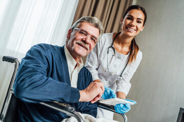 An older man in a wheelchair, next to him his female doctor shows something in the tablet. They smile happily and look at the camera