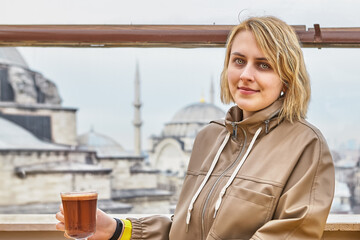 Beautiful European young woman in her 20s with glass of tea by window in cafe overlooking Istanbul cityscape looking at camera.