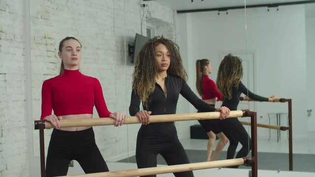 Motivated sporty fit black female exercising plie squat at training barre, strengthening leg and hip muscles while slim professional woman fitness trainer correcting her posture in fitness studio.