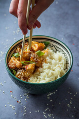 Woman eating sweet and sour sauce chicken using chopsticks. Traditional Asian street food - Crispy sesame chicken with rice in a green decorated bowl. Homemade dish, ready to eat. Gray background