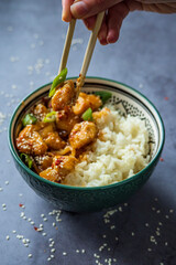 Hand eating with chopsticks traditional Asian street food - Crispy sesame chicken in sweet and sour sauce with portion of rice in a green decorated bowl. Homemade dish, ready to eat. Gray background