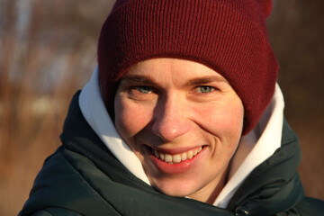 A blue-eyed girl in a burgundy knitted hat and a white hood smiles against the background of a spring forest.