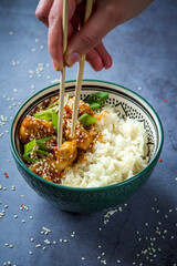 Woman eating sweet and sour sauce chicken using chopsticks. Traditional Asian street food - Crispy sesame chicken with rice in a green decorated bowl. Homemade dish, ready to eat. Gray background