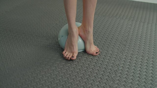 Closeup of slim barefoot muscular female legs standing on medicine ball and practicing squat, exercise for balance and stability,. Low section of fit woman working out in gym using fitness mini ball.
