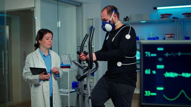 Performance athlete talking with medical researcher running on on cross trainer in sports science laboratory measuring VO2 max, heart rate, psychological resistance and muscular endurance. Monitor