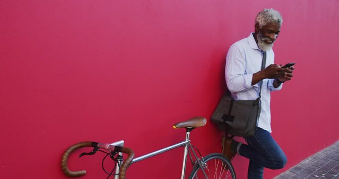 African american senior man with bicycle using smartphone while standing against pink wall