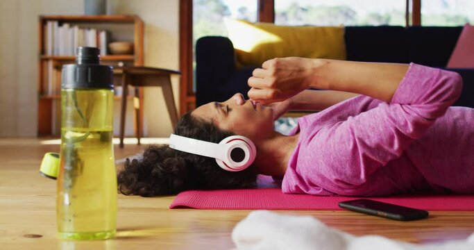 Mixed race woman wearing headphones practicing yoga and meditating while lying on yoga mat at home