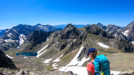 A young woman with a big backpack hikes down towards a clear, navy blue lake hiding between tall...