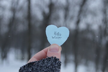 Hello winter. Hand holds a blue heart with the inscription on the background of the winter forest.