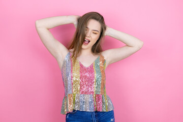 Pretty redhead woman wearing a shiny top standing over pink background relaxing and stretching, arms and hands behind head and neck smiling happy