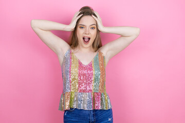 Pretty redhead woman wearing a shiny top standing over pink background crazy and scared with hands on head, afraid and surprised of shock