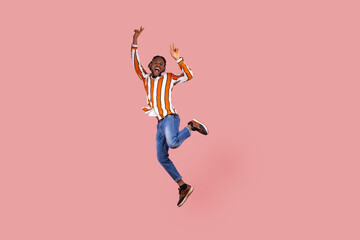 Fototapeta na wymiar Full length excited ecstatic afro-american man with dreadlocks in striped shirt highly jumping posing, showing victory gesture, celebrating his triumph. Indoor studio shot isolated on pink background