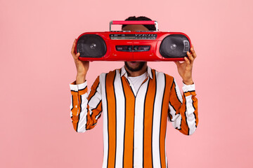 Funny afro-american man with beard and dreadlocks in bright striped shirt hiding his face behind red vintage audio record player, selling junk. Indoor studio shot isolated on pink background
