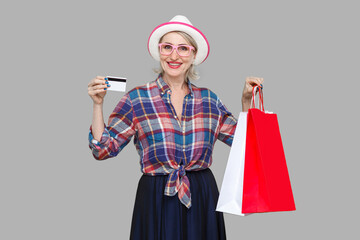 Funny aged woman in white hat and pink eyeglasses, in checkered shirt standing, holding bank credit card and shopping bags with toothy smile, looking at camera. Indoor, studio shot, gray background