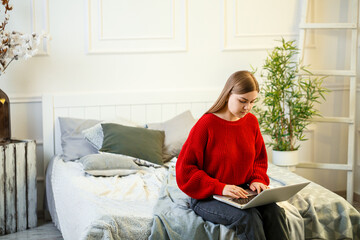 Young woman working on laptop sitting on the bed at home. Work from home during quarantine. Girl in a sweater and jeans at home on the bed