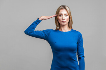 Yes sir! Portrait of serious woman in elegant tight blue dress standing saluting with hand near head, looking obedient and attentive at camera. indoor studio shot isolated on gray background