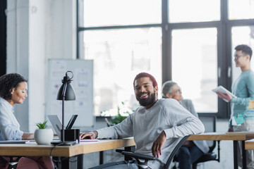 Naklejka na ściany i meble Smiling african american businessman sitting near laptop in office