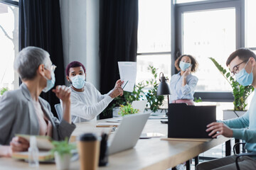 African american businessman in medical mask pointing at paper near multiethnic colleagues