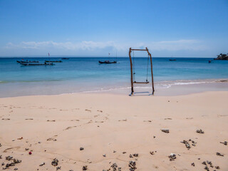 A swing placed on the seashore of Pink Beach, Lombok, Indonesia. The swing has very simple wood construction. Waves gently wash the pillars of it. In the back there are few boats anchored in the bay.