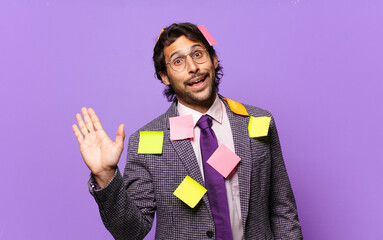 young handsome indian man smiling happily and cheerfully, waving hand, welcoming and greeting you, or saying goodbye. humorous business concept