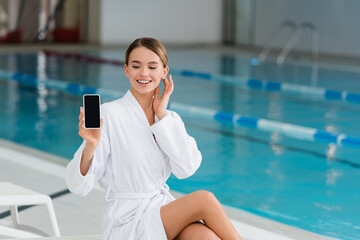 cheerful woman in white bathrobe holding smartphone with blank screen in spa center