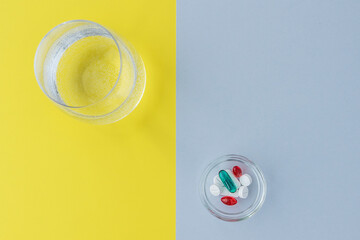 top view of little round plate with pills of different shapes and colors and glass of water on background of colors of 2021, grey and yellow. Healthcare concept
