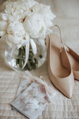 Fototapeta na wymiar Wedding bouquet of peonies flowers in a vase stands on the bed of the newlyweds with invitations and shoes on the background of the bride dresses