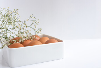 Easter eggs in a white square dish, on a white table, with a sprig of delicate flowers. Close-up, space for text
