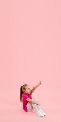Pointing. Childhood and dream about big and famous future. Pretty little girl isolated on coral pink studio background. Dreams, imagination, education, facial expression, emotions concept. Flyer