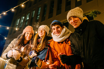 Group of four people student in warm clothes with burning sparklers or bengal lights enjoying parety in winter outdoors.